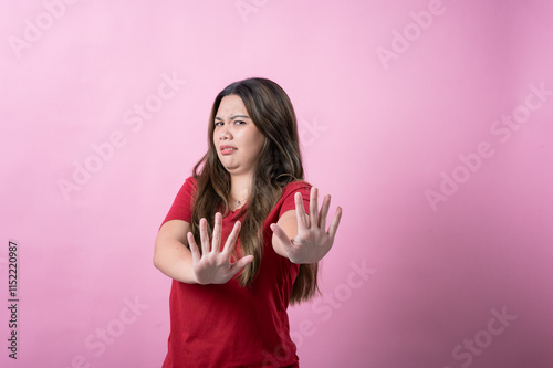 A young woman with long brown hair and light skin, wearing a red T-shirt, makes a disgusted expression while raising her hands defensively, standing against a solid pink background.