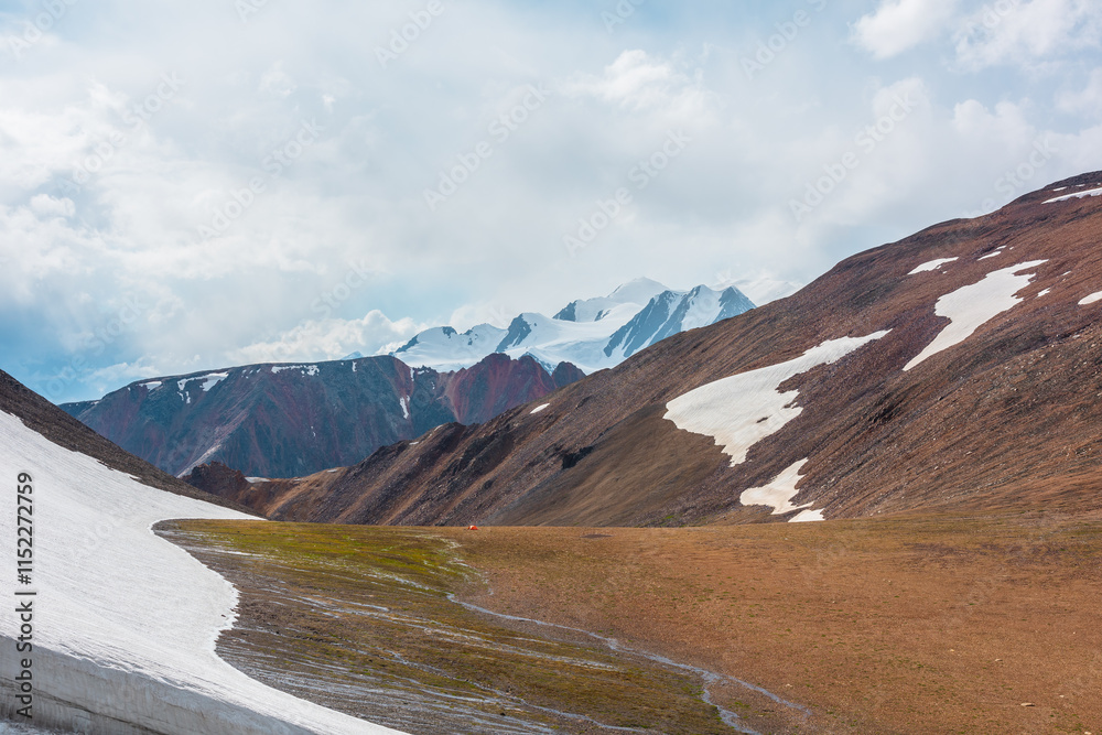 Φωτογραφία Orange tent near glacier in sunlit high alpine valley against big red rocky ridge and large snowy peak far away in low clouds