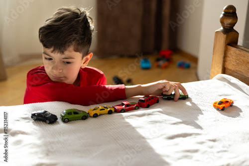 A young boy, aged 6-8, plays with toy cars in a sunny, cozy room, aligning them in a row. The scene highlights themes of autism, developmental disorders, childhood play, and mental well-being. photo