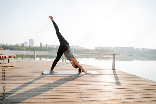A woman is practicing yoga on a beautiful dock that is situated close to a serene lake, enjoying the peaceful surroundings and the calming atmosphere while engaging in physical fitness activities