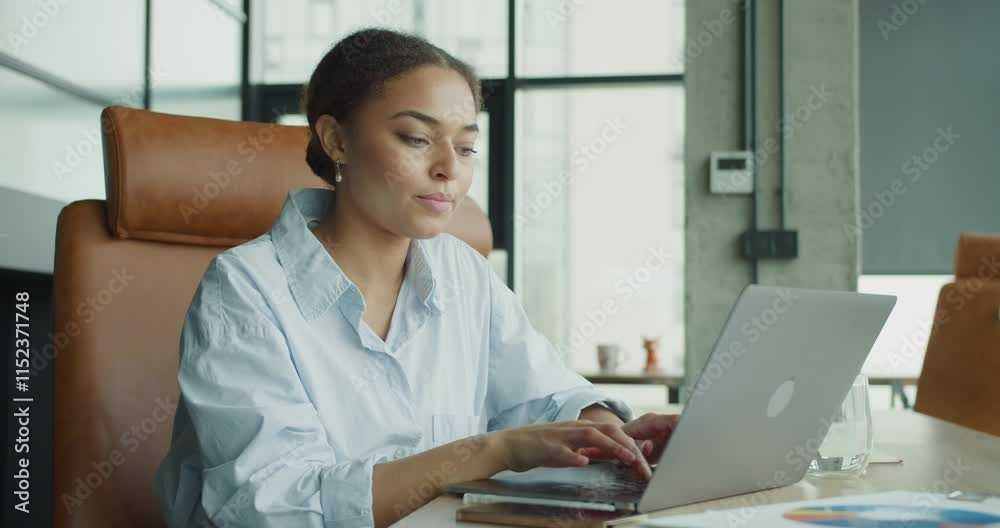A focused young woman working on a laptop in a modern office environment, interacting with a smartphone and analyzing documents on a desk with bright natural lighting