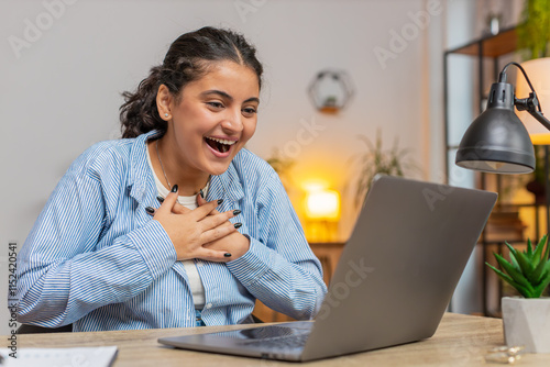 Happy young Indian woman working on laptop netbook shocked by sudden victory, winning, goal achievement, good career news. Excited freelancer girl clenching fists at home office workplace. Lifestyle. photo