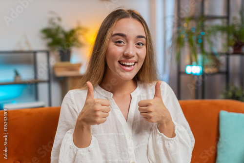 Portrait of smiling Caucasian woman looking approvingly at camera showing double thumbs up, like sign, positive feedback, good news. Happy girl sitting on couch sofa in living room at home. Lifestyle photo