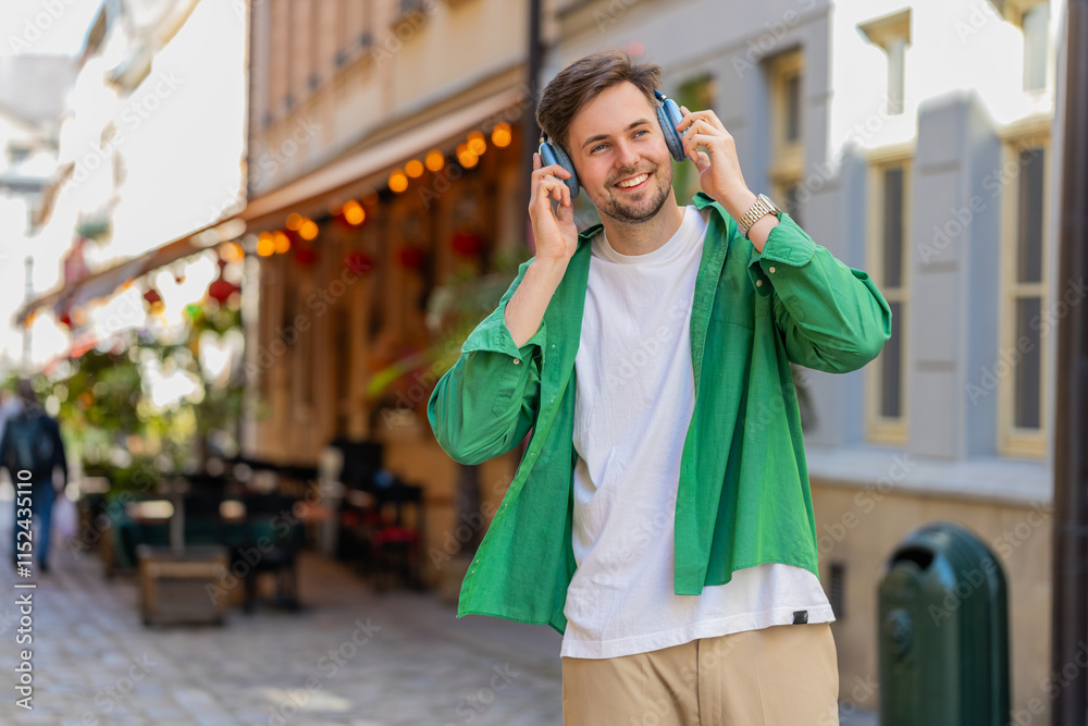 Fototapeta premium Handsome young man tourist in wireless headphones listening favorite energetic disco music outdoors. Happy Caucasian male traveler walking passes by urban sunny city street. Town lifestyles