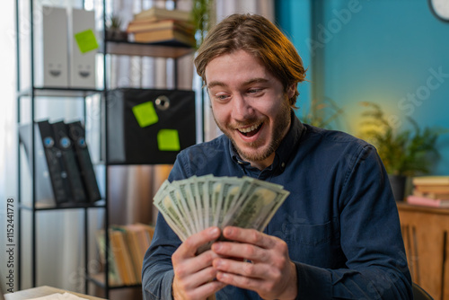Excited businessman guy holding fan of cash money dollar banknotes showing thumbs up and pointing at laptop, celebrate, success career, online income, wealth at home office. Remote distant working.