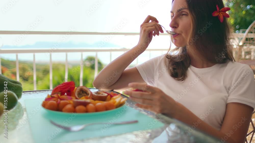 Beautiful woman is enjoying a delicious plate of exotic fruits while sitting on a balcony overlooking a tropical landscape