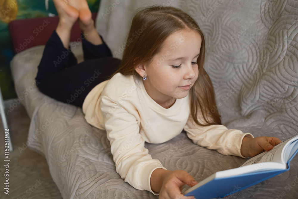 A young girl is lying on a couch, comfortably reading a book in a cozy atmosphere. The image portrays leisure, focus, and the joy of reading in a calm and peaceful environment.