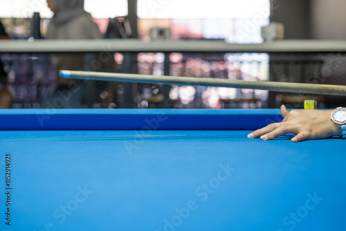 A hand strikes a billiard ball with precision using a cue stick during an intense pool game on a blue table, highlighting focus, skill, and competitive gameplay in an indoor setting. photo