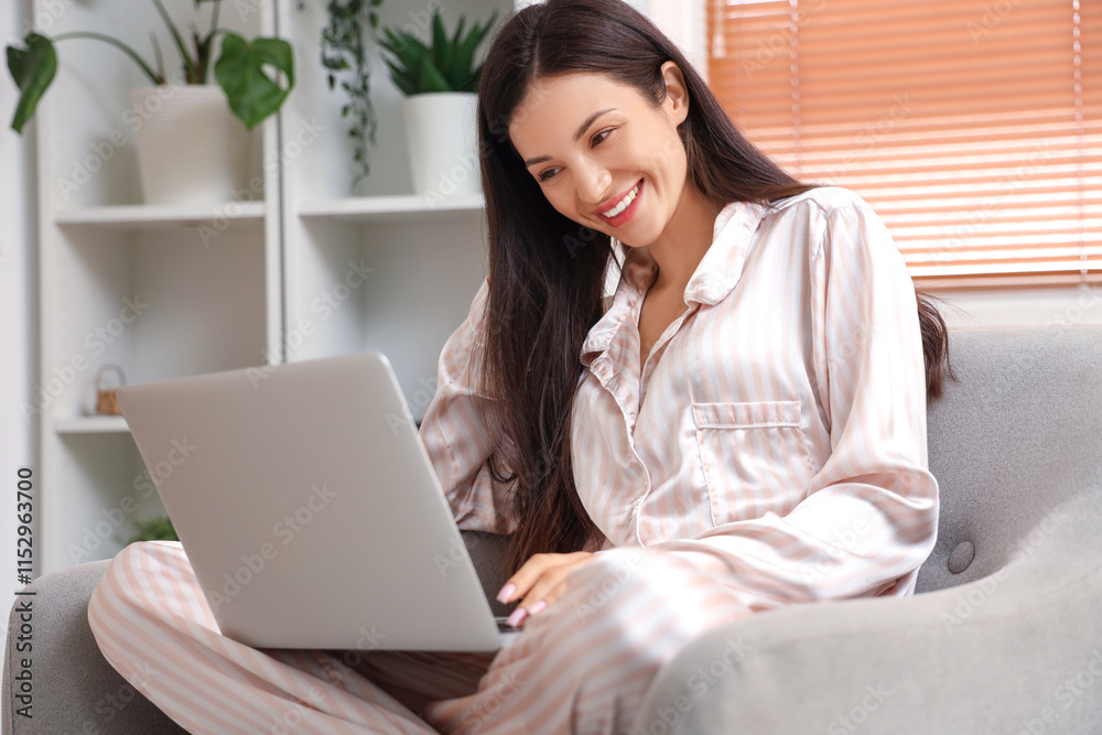 Female freelancer in striped pajamas working with laptop on armchair at home Female freelancer in striped pajamas working with laptop on armchair at home