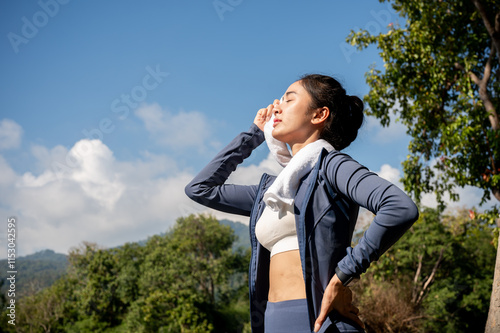 Tired, sweaty Asian woman in sportswear wipes sweat from her face with a towel, exercising in a park