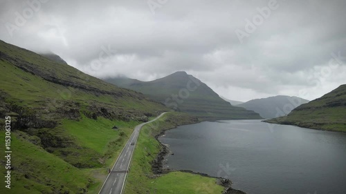 A scenic coastal road in Funnings, Faroe Islands, winds through lush green landscapes and rugged cliffs, with calm waters and dramatic peaks under a cloudy sky, offering a tranquil and remote view photo