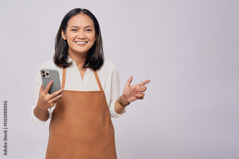 Smiling young Asian woman barista barman employee wearing a brown apron working in coffee shop holding mobile phone and pointing finger at copy space isolated on white background Smiling young Asian woman barista barman employee wearing a brown apron working in coffee shop holding mobile phone and pointing finger at copy space isolated on white background