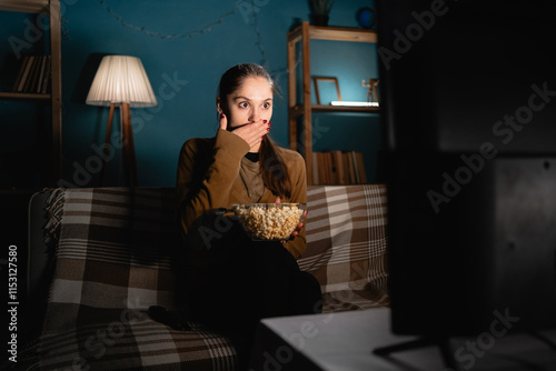 Shocked woman watching scary movie covers mouth in shock sitting near tv eating popcorn in living room at home at night. photo