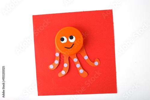 Orange plastic bottle cap octopus with paper tentacles and dotted details on a red square paper. Minimalist flat lay, Craft and DIY recycling concept. preschool hour teaching activity, photo