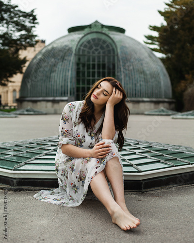 A woman sits barefoot, cross-legged, and rests her hand on her head, closing her eyes. In the background is a large dome-shaped glass structure, reminiscent of a greenhouse or botanical garden. photo