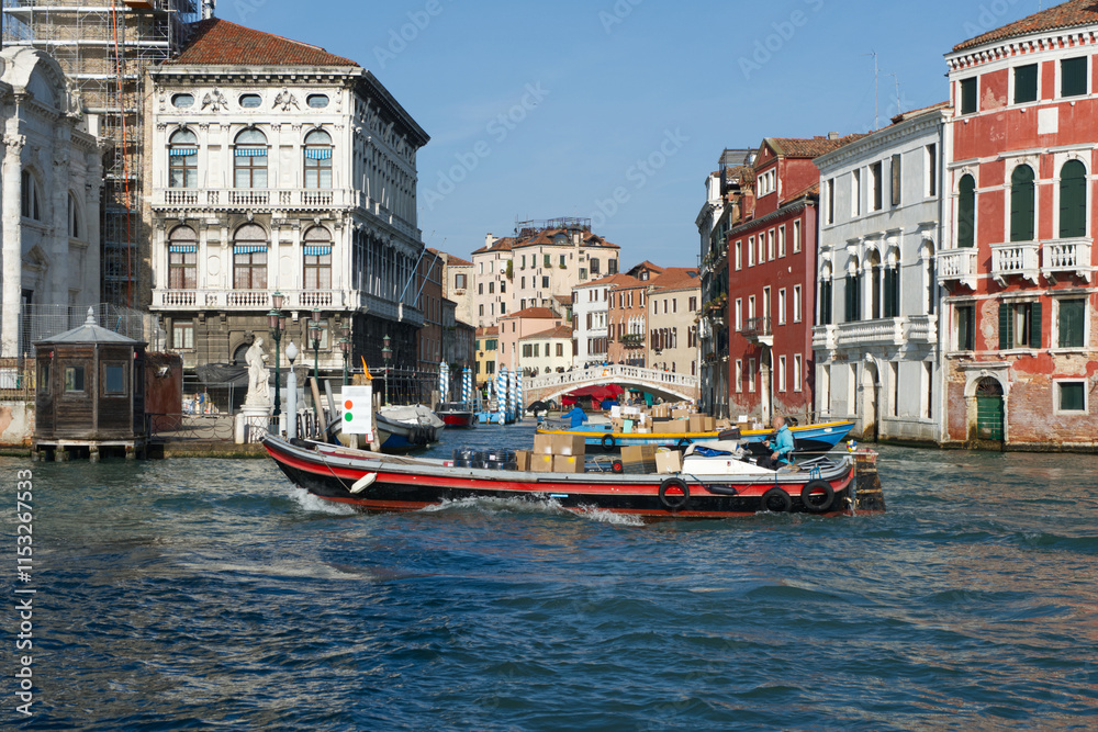 Obraz premium A boat carrying packages navigates a Venetian canal, surrounded by historic buildings and a bridge, showcasing the city's unique waterways and architecture.