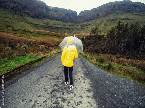 Travel and tourism in Ireland. Teenager girl with umbrella on a road and looking at majestic nature scenery of Gleniff Horseshoe drive with tall mountains. Model wearing yellow jacket and hat. photo