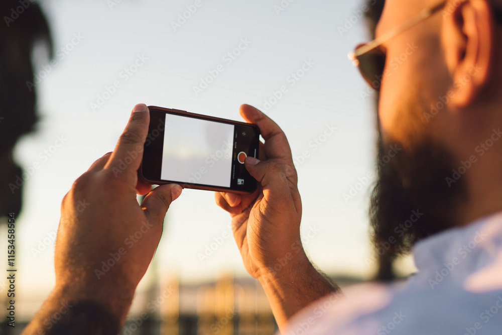 Back view of male blogger using smartphone with mock up screen for taking pictures and share in networks, hipster guy holding modern mobile phone making photos during summer vacations in evening Back view of male blogger using smartphone with mock up screen for taking pictures and share in networks, hipster guy holding modern mobile phone making photos during summer vacations in evening