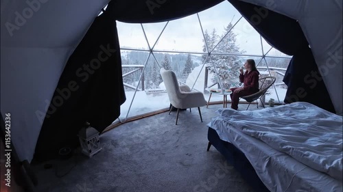 Woman in pajamas eating breakfast in dome hotel near window with view of mountains and snow.  Luxury glamping tent. Cozy hygge travel holidays retreat destination
