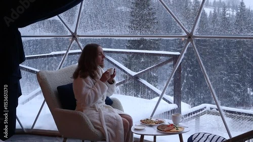 Woman sits near a window in a dome hotel in the mountains in winter and drinking coffee. Luxury glamping tent. Cozy hygge travel holidays retreat destination
