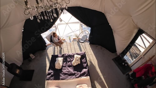 Woman sits near a window in a dome hotel in the mountains in winter and reading book. Luxury glamping tent. Cozy hygge travel holidays retreat destination
