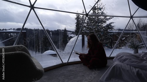 Woman sits near a window in a dome hotel in the mountains in winter and reading book. Luxury glamping tent. Cozy hygge travel holidays retreat destination
