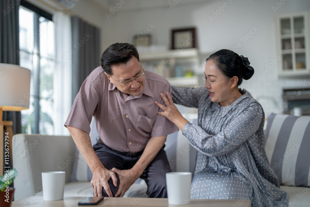 An elderly couple sits in their living room. The man holds his knee, appearing to be in discomfort, while his partner offers support. The image conveys care, concern, and the bond between the couple. An elderly couple sits in their living room. The man holds his knee, appearing to be in discomfort, while his partner offers support. The image conveys care, concern, and the bond between the couple.