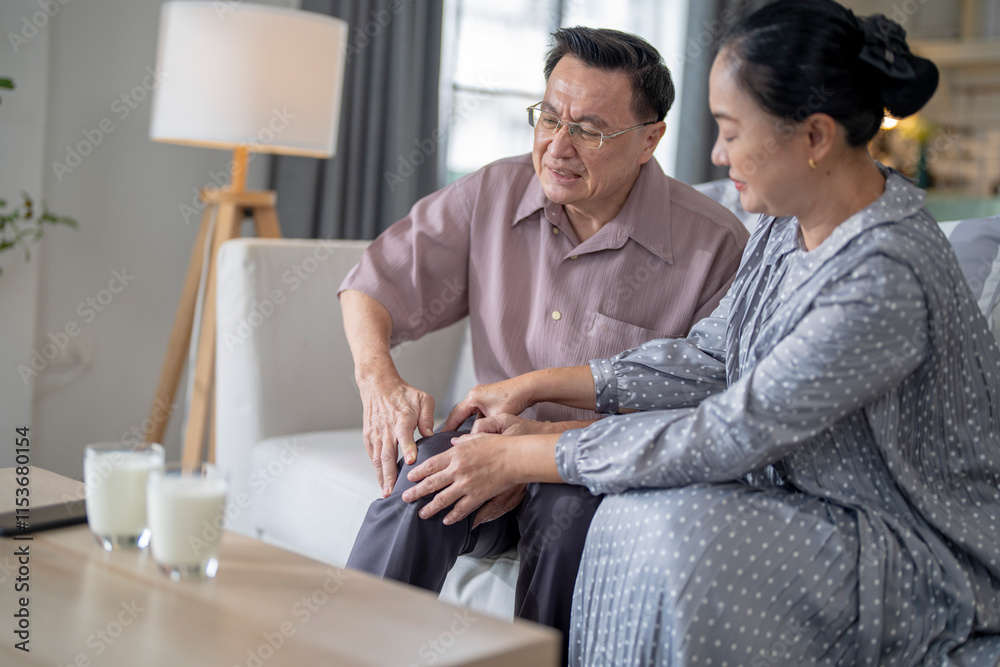An elderly couple sits in their living room. The man holds his knee, appearing to be in discomfort, while his partner offers support. The image conveys care, concern, and the bond between the couple.