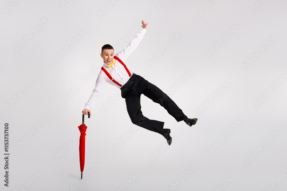 Fototapeta premium Young man leaps joyfully with vibrant red umbrella against white background, embodying dance and fashion
