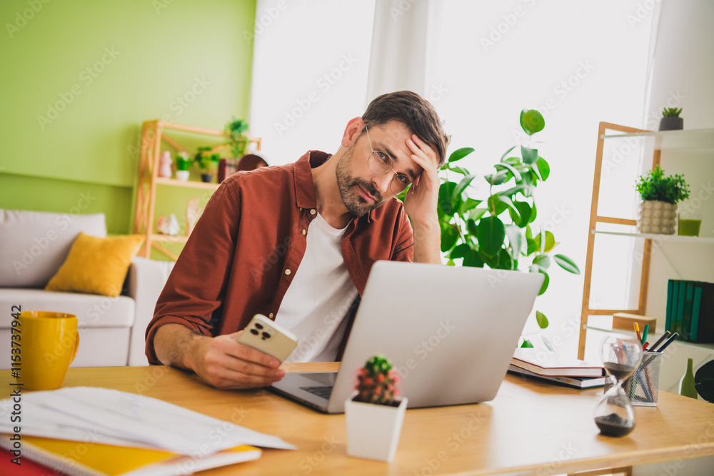 Man holding phone and sitting at desk with laptop in bright home interior Man holding phone and sitting at desk with laptop in bright home interior
