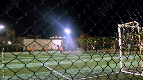 Handheld real-time footage of unrecognizable men playing soccer. Night football field as seen from outside