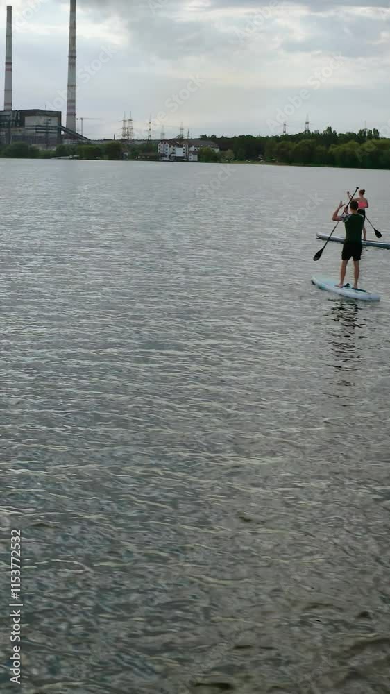 Young people sailing on water. Man and woman travelling on river on paddle boards on industry background in the evening. Summer rest. Vertical video Young people sailing on water. Man and woman travelling on river on paddle boards on industry background in the evening. Summer rest. Vertical video