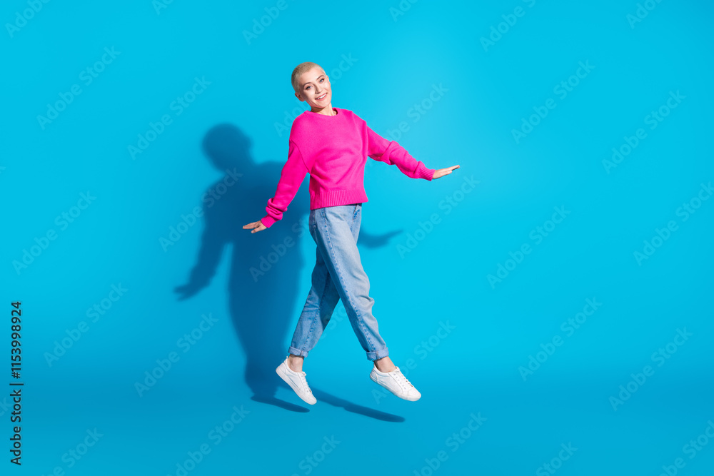 Obraz premium Joyful young woman jumping in mid-air against a vibrant blue background, wearing a bright pink sweater and jeans