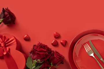 Valentine's day. Romantic table setting with white plate, red chocolate sweets, romantic gift and red roses on red background. Catering. View from above. Copy space.