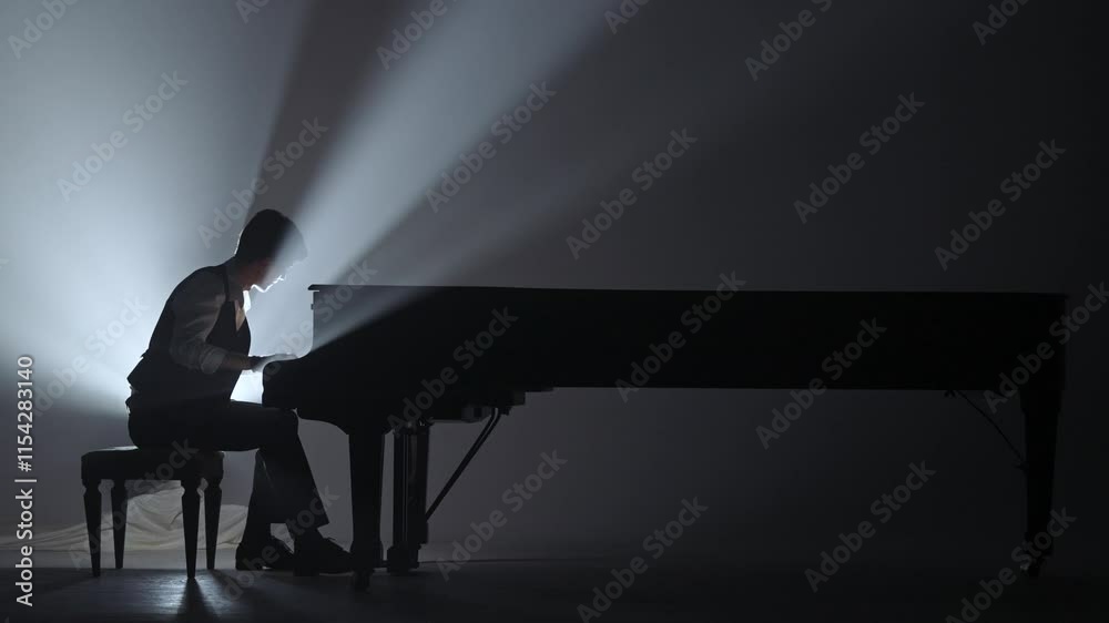 Pianist sitting and playing on the black grand piano on dark concert stage against bright spotlight, isolated on black studio background. Artist performing in concert hall.
