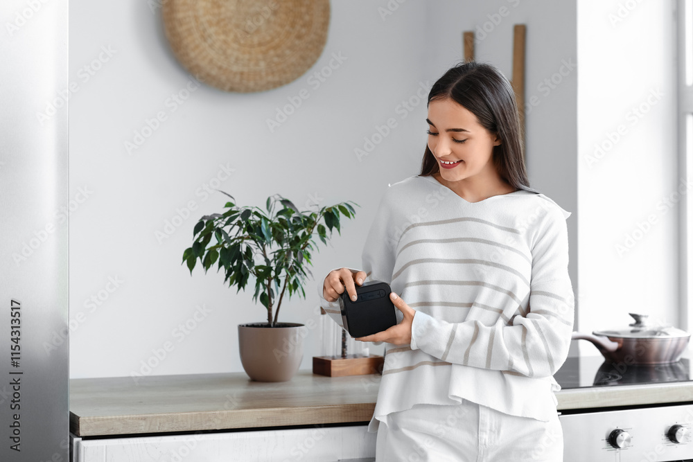 © Pixel-Shot - Beautiful young happy woman with wireless portable speaker in kitchen at home