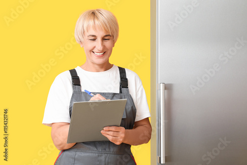 Female worker with clipboard near refrigerator on yellow background