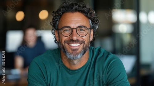 A happy man with curly hair and a beard smiles while wearing glasses and a green shirt, captured in a stylish indoor office space with a blurred background.