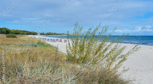 Fototapeta Naklejka Na Ścianę i Meble -  A peaceful view of the Baltic Sea coastline with grassy dunes, strandkorb beach chairs, and calm blue waters under a partly cloudy sky. A serene natural landscape.