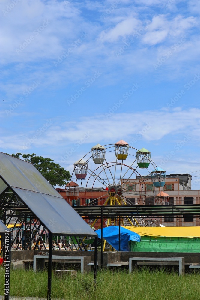 Several small, covered stalls are set up in a grassy field with a Ferris wheel in the background. Several small, covered stalls are set up in a grassy field with a Ferris wheel in the background.