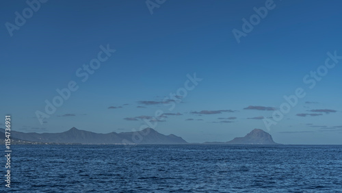 The coast of a tropical island is visible from the ocean. Picturesque silhouettes of mountains against the background of blue sky and clouds. Ripples on the surface of the water. Mauritius. Copy Space