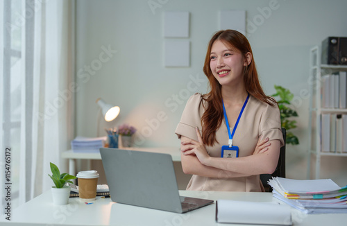 A beautiful young woman, a confident professional, is happily sitting with her arms crossed at her desk with a laptop and a pile of paperwork. Smile beautifully and charmingly in a modern office. photo