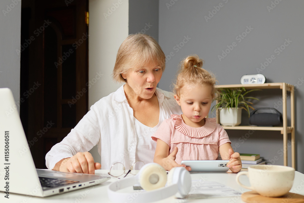 A grandmother and her granddaughter sit together at a table, focused on a digital device while surrounded by a laptop and a cup. This intimate moment captures their shared enjoyment and curiosity