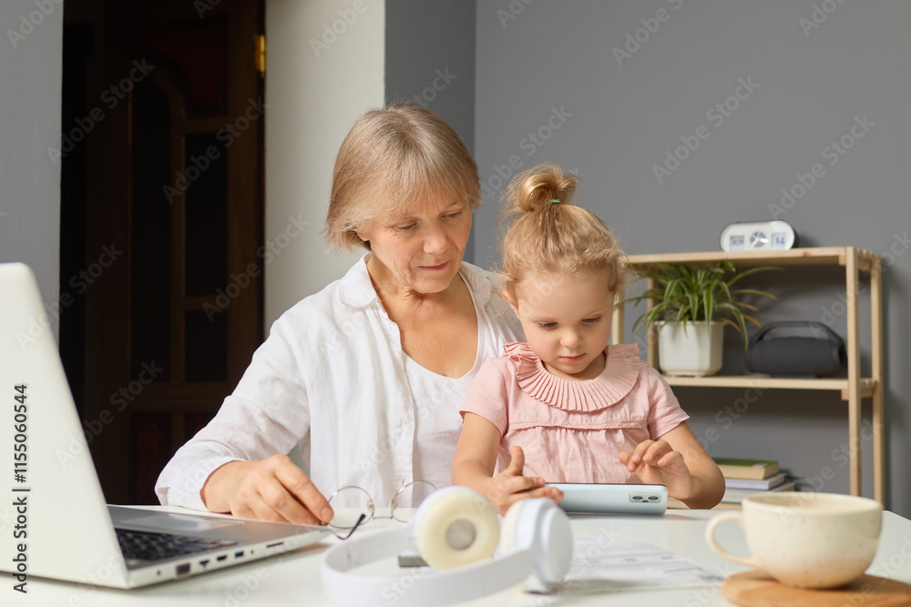 A grandmother and her young granddaughter sit at a table, engaged in a shared activity. The girl interacts with a device while her grandmother observes with joy, creating a warm family moment A grandmother and her young granddaughter sit at a table, engaged in a shared activity. The girl interacts with a device while her grandmother observes with joy, creating a warm family moment