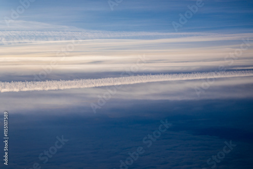 Dramatic contrail stretches across layered clouds in high altitude aerial view. Natural abstract pattern of aircraft vapour trail against deep blue sky and stratiform cloud formations