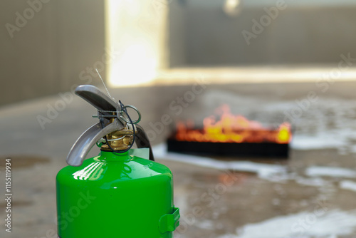 A green fire extinguisher stands in focus, while a controlled fire burns in the background, highlighting fire safety equipment in use. photo