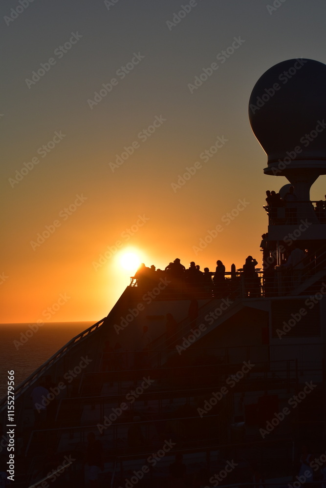 Obraz premium A stunning sunset over the ocean with silhouettes of passengers on a ship's deck, the warm glow of the sun highlighting the peaceful scene as it sets on the horizon, creating a serene atmosphere at se