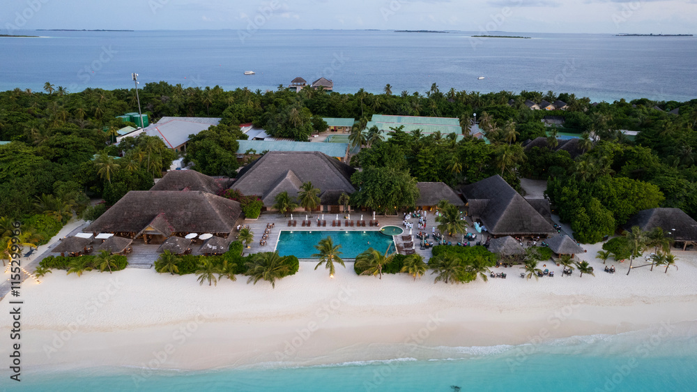White sand on the beach and a large swimming pool. Aerial view from a drone of an island in the ocean, turquoise clear water. Green palm trees all over the island. Houses with thatched roofs. Rest