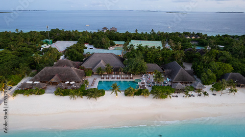 Obraz na płótnie White sand on the beach and a large swimming pool. Aerial view from a drone of an island in the ocean, turquoise clear water. Green palm trees all over the island. Houses with thatched roofs. Rest w salonie