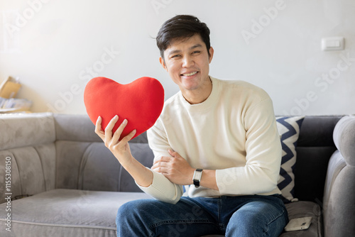 close up young asian man smile with happiness feeling and hold red color of heart shape pillow while sit on couch in living room for good health and live better and valentine day and people lifestyle photo